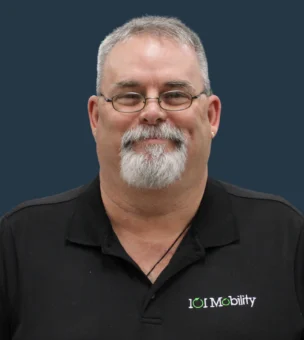 Man with glasses and a beard wearing a black polo shirt with a "101 Mobility" logo, standing against a dark background.
