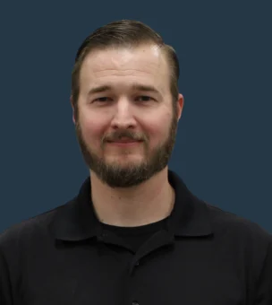 A person with short hair and a beard is wearing a black collared shirt, standing in front of a plain dark blue background.