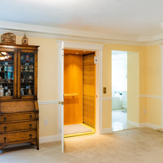 A wooden cabinet stands next to an open home elevator door in a warmly lit room with beige walls and carpet flooring.