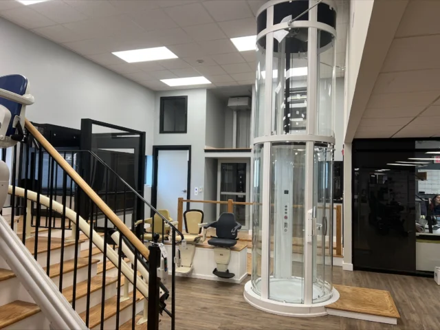 Modern interior with a cylindrical glass elevator beside a staircase equipped with two stairlifts. The area has wood flooring and bright lighting.