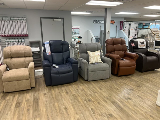 Four recliners in different colors are displayed in a showroom with wooden flooring and shelves in the background.