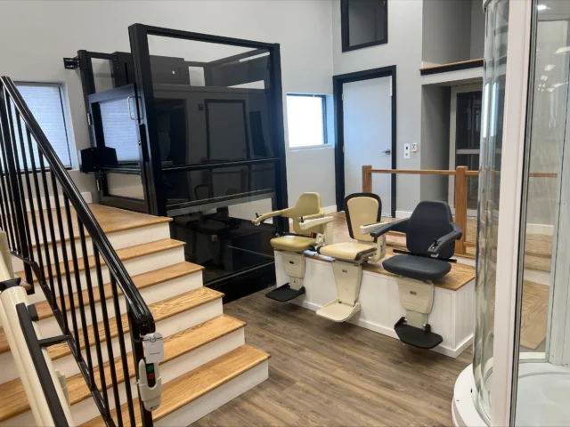 A showroom displaying a home elevator, a curved stairlift on a wooden staircase, and three seated stairlifts on a platform.