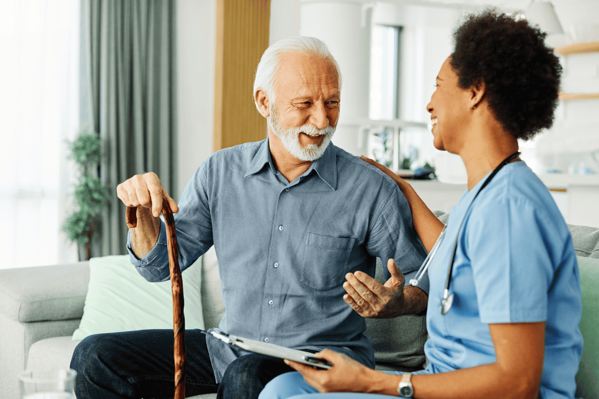 A smiling older man with a cane sits on a couch, talking to a healthcare professional in scrubs.