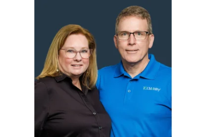 Two people posing together against a dark background. One is wearing a black shirt, and the other is in a blue polo.