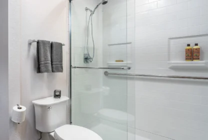 Bathroom with a glass-enclosed shower, white tiled walls, toilet, toilet paper holder, towel rack with gray towels, and two bottles on a shower shelf.