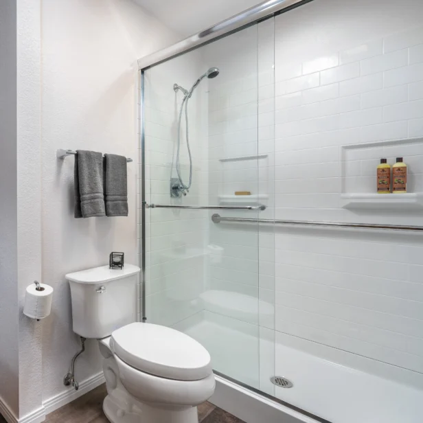 Bathroom with a glass-enclosed shower, white tiled walls, toilet, toilet paper holder, towel rack with gray towels, and two bottles on a shower shelf.