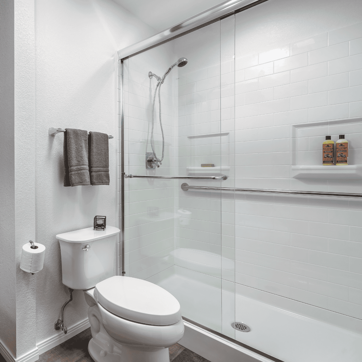 Bathroom with a glass-enclosed shower, white tiled walls, toilet, toilet paper holder, towel rack with gray towels, and two bottles on a shower shelf.