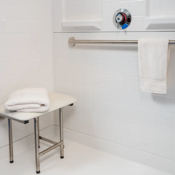 Accessible shower with a safety grab bar, control knobs, and a foldable shower chair. Towels are placed on the chair and on the grab bar.