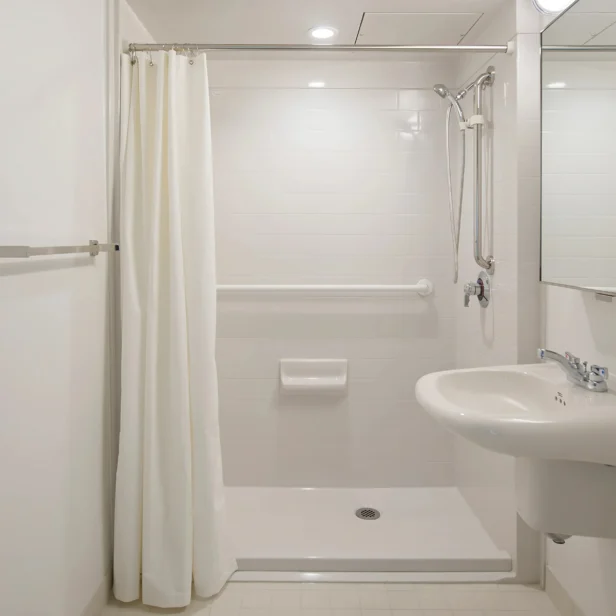 White bathroom with a shower, curtain, grab bar, showerhead, wall-mounted soap dish, sink with faucet, and mirror. Clean, minimal design.