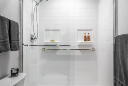 A modern shower with glass doors, white tiles, and built-in shelves holding toiletries. Towels hang on the wall, and a toilet is visible in the foreground.