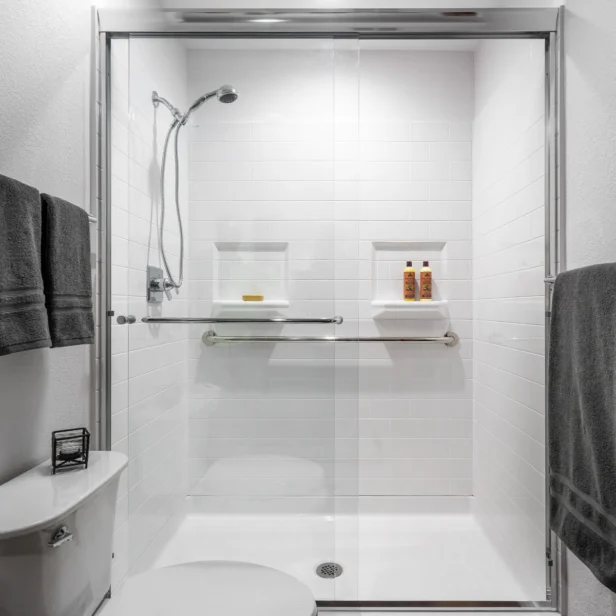 A modern shower with glass doors, white tiles, and built-in shelves holding toiletries. Towels hang on the wall, and a toilet is visible in the foreground.