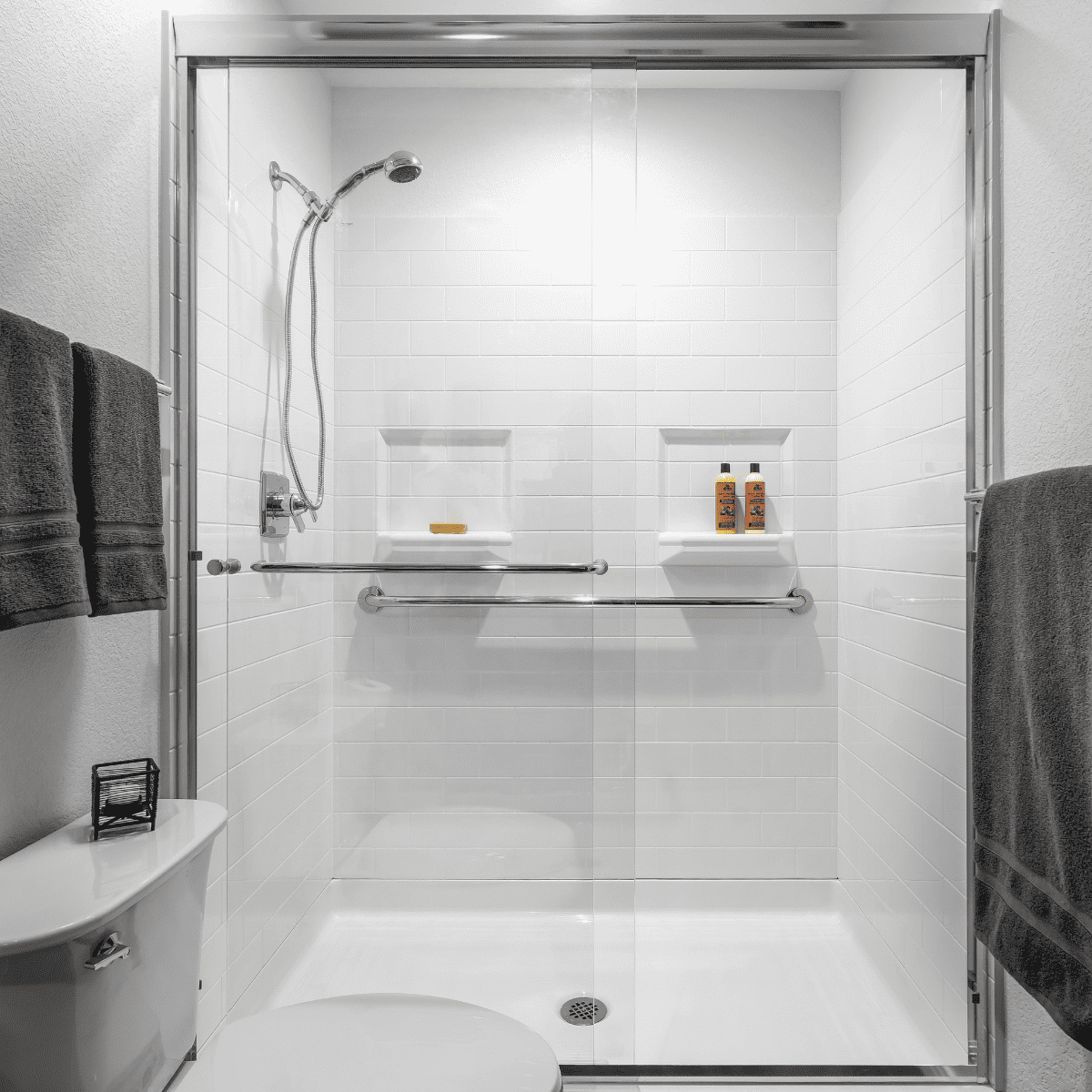 A modern shower with glass doors, white tiles, and built-in shelves holding toiletries. Towels hang on the wall, and a toilet is visible in the foreground.