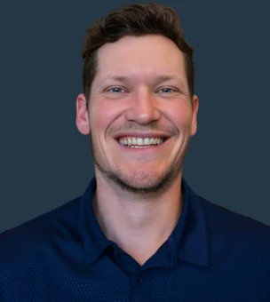 Smiling person in a dark blue collared shirt against a plain dark background.