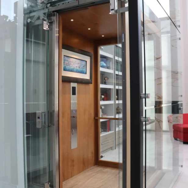 Modern glass elevator with wooden interior, spotlighting, and a framed picture. Nearby, a white bookshelf with various items and a red chair are visible in a bright room.