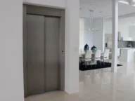 A modern indoor space with an elevator on the left, a dining area with a white table and chairs in the middle, and a kitchen with sleek appliances on the right.