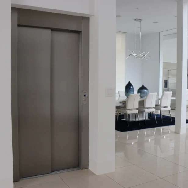 A modern indoor space with an elevator on the left, a dining area with a white table and chairs in the middle, and a kitchen with sleek appliances on the right.