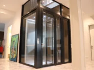 Modern glass and metal elevator inside a home, with a painting and a red toy nearby on the tiled floor.