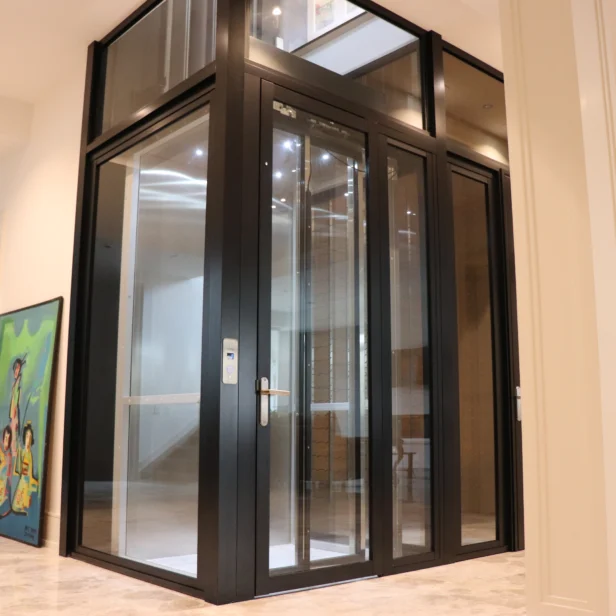 Modern glass and metal elevator inside a home, with a painting and a red toy nearby on the tiled floor.