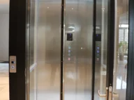 Glass elevator with open doors, showing control panels and reflective interior. Elevator is situated in a modern building with marble flooring.