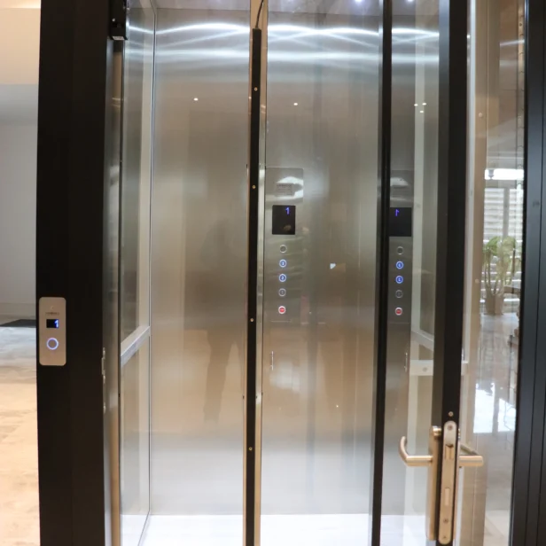 Glass elevator with open doors, showing control panels and reflective interior. Elevator is situated in a modern building with marble flooring.
