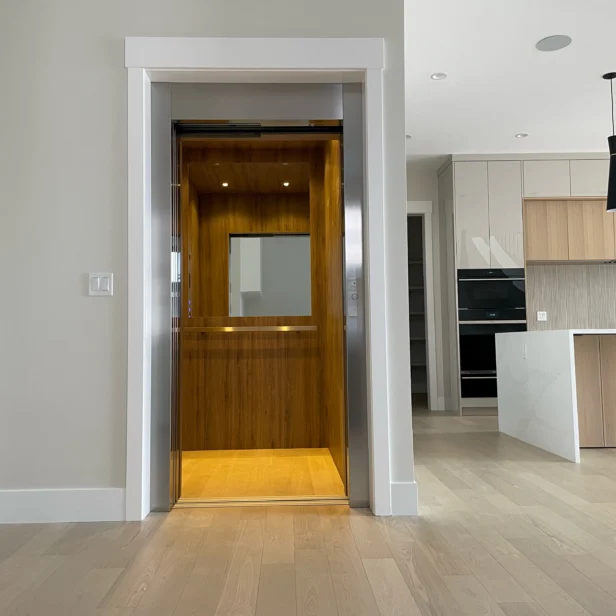 A modern home interior featuring an open elevator with wood paneling. Adjacent is a kitchen area with light wood cabinets, a black pendant light, and a marble island.