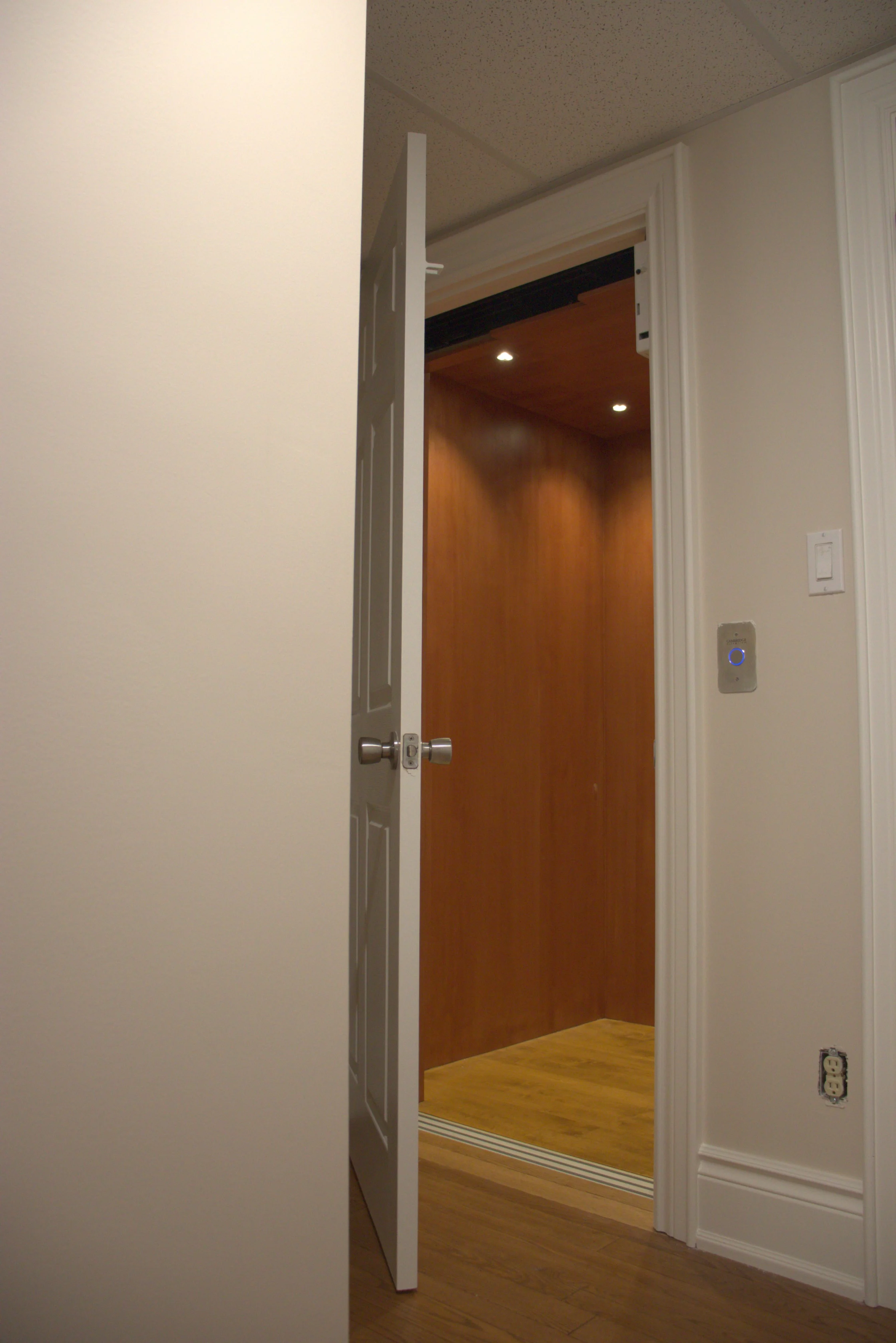 Open door revealing an interior residential elevator with wood paneling and recessed lighting.