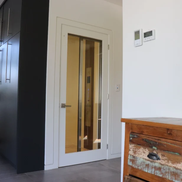 Interior with a modern, glass-paneled elevator door, dark cabinetry to the left, a wooden dresser to the right, and a staircase leading upwards.