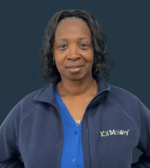 A person with shoulder-length hair wearing a blue shirt and a navy jacket with a logo, standing against a plain dark background.