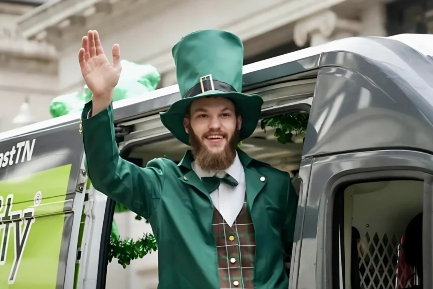 Person in leprechaun costume with a green top hat and plaid vest waves from a vehicle window.