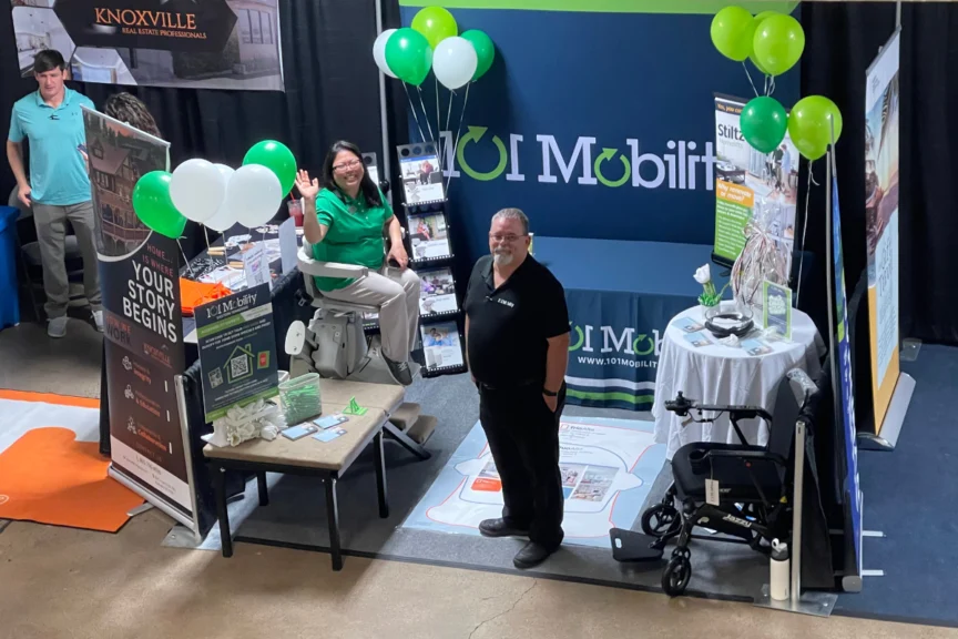 Exhibit booth showcasing mobility solutions with two individuals, brochures, a chair lift, and balloons.