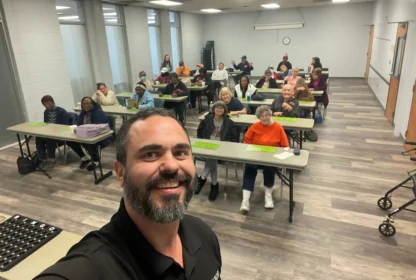 A man takes a selfie at the front of a classroom filled with people seated at tables.