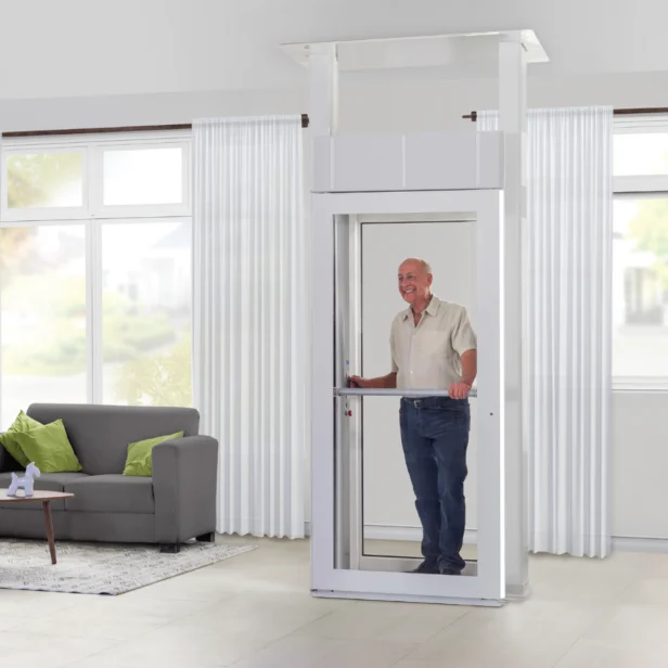 An elderly man stands inside a white home elevator in a living room with a gray sofa and green cushions.