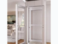 Glass-enclosed home elevator with an open door, leading to a living room with a couch and painting on the wall.