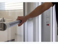 Person holding an elevator handrail beside a control panel. A couch is visible through the glass door.