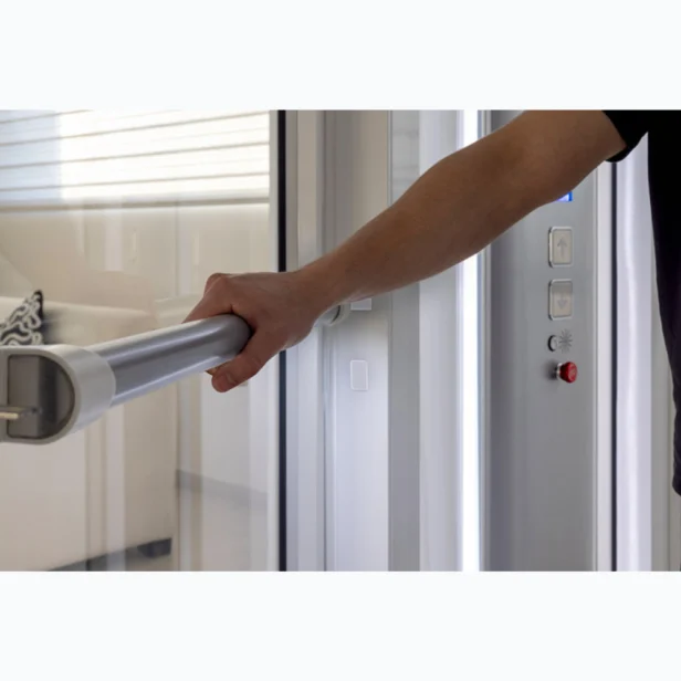 Person holding an elevator handrail beside a control panel. A couch is visible through the glass door.