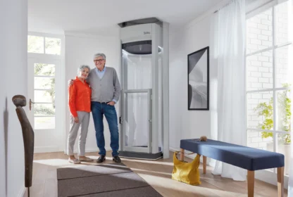 An elderly couple stands beside a modern home elevator in a bright, well-lit room with large windows, a bench, and a decorative sculpture.