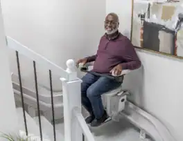 A man sits on a motorized stair lift chair, ascending a curved staircase in a modern, well-lit home.