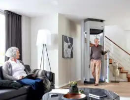 An older man exits a residential elevator while a woman sits on a sofa reading in a modern living room with a staircase in the background.