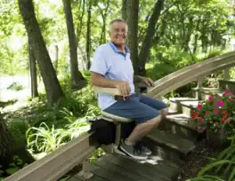 An older man rides an outdoor stair lift surrounded by greenery and flowers on a sunny day.