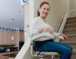 A woman sits on a stair lift attached to a staircase in a home, smiling at the camera. Kitchen cabinets and appliances are visible in the background.
