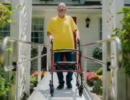 An older man in a yellow shirt stands with a walker on a ramp outside a house with a garden and white railing.