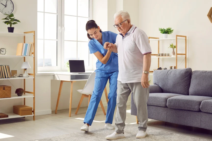 A nurse in blue scrubs assists an elderly man with glasses as he walks in a bright living room with modern furniture.