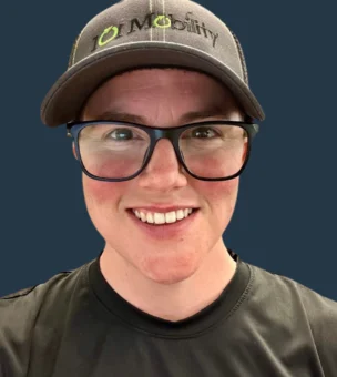 Person wearing black-rimmed glasses, a black "IO Mobility" hat, and a black shirt, smiling at the camera with a plain dark blue background.