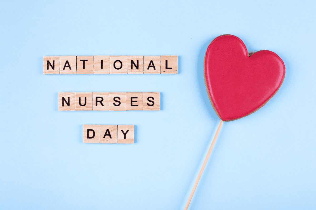 Scrabble tiles spell "National Nurses Day" next to a red heart-shaped object on a stick, all set against a light blue background.