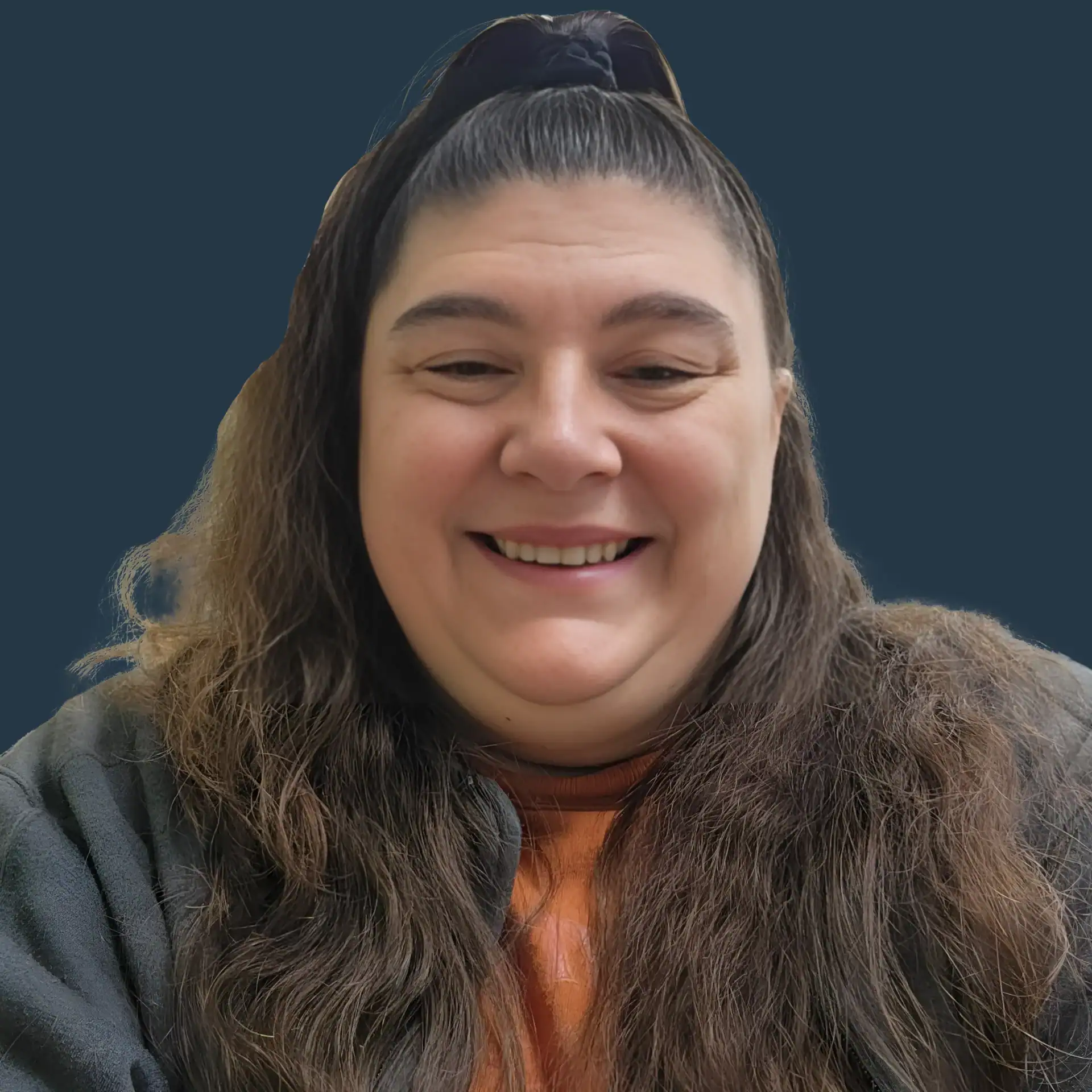 A person with long brown hair and a ponytail smiles at the camera, wearing a dark jacket and orange shirt, with a plain dark blue background.