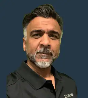 A man with short, dark hair and a salt-and-pepper beard wearing a black collared shirt, standing against a plain dark background.