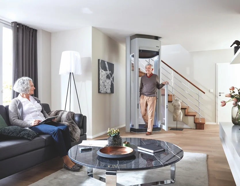 An elderly man exits a home elevator while an elderly woman sits on a sofa reading in a modern living room.