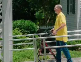 A man in a yellow shirt uses a walker while walking up a ramp outside a white house on a sunny day.