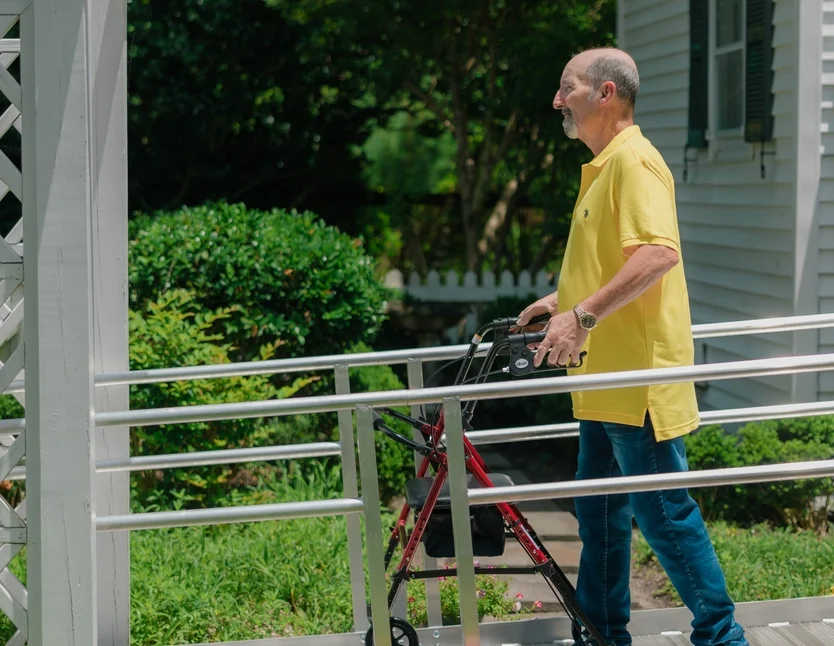 A man in a yellow shirt uses a walker to move along an outdoor wheelchair-accessible ramp near a white building and green shrubs.