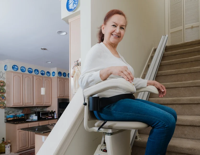 A woman sits and smiles on a stair lift chair attached to the staircase in a home kitchen area.